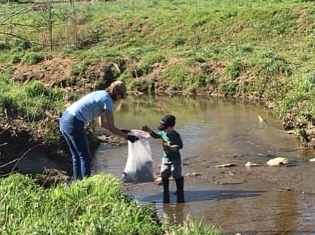 A woman holding out a trash bag to a boy standing in a creek picking up trash.