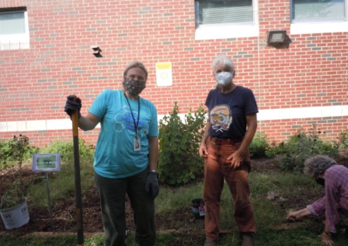 Two volunteers at a planting holding their shovels while another volunteer installs plants in the background.