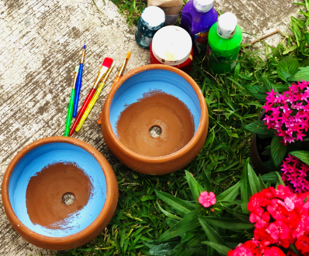 Two clay flower pots with blue paint on them sitting beside paintbrushes and bottles of paint.