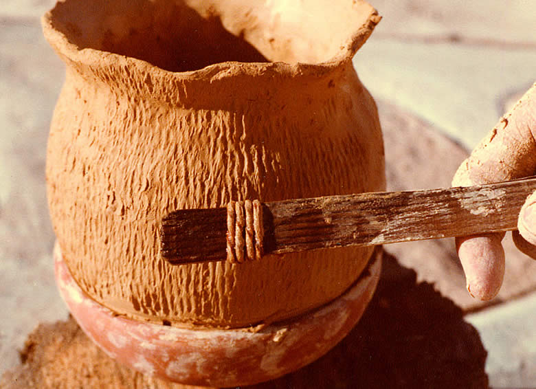 An artist adds texture to a clay pot with a tool.